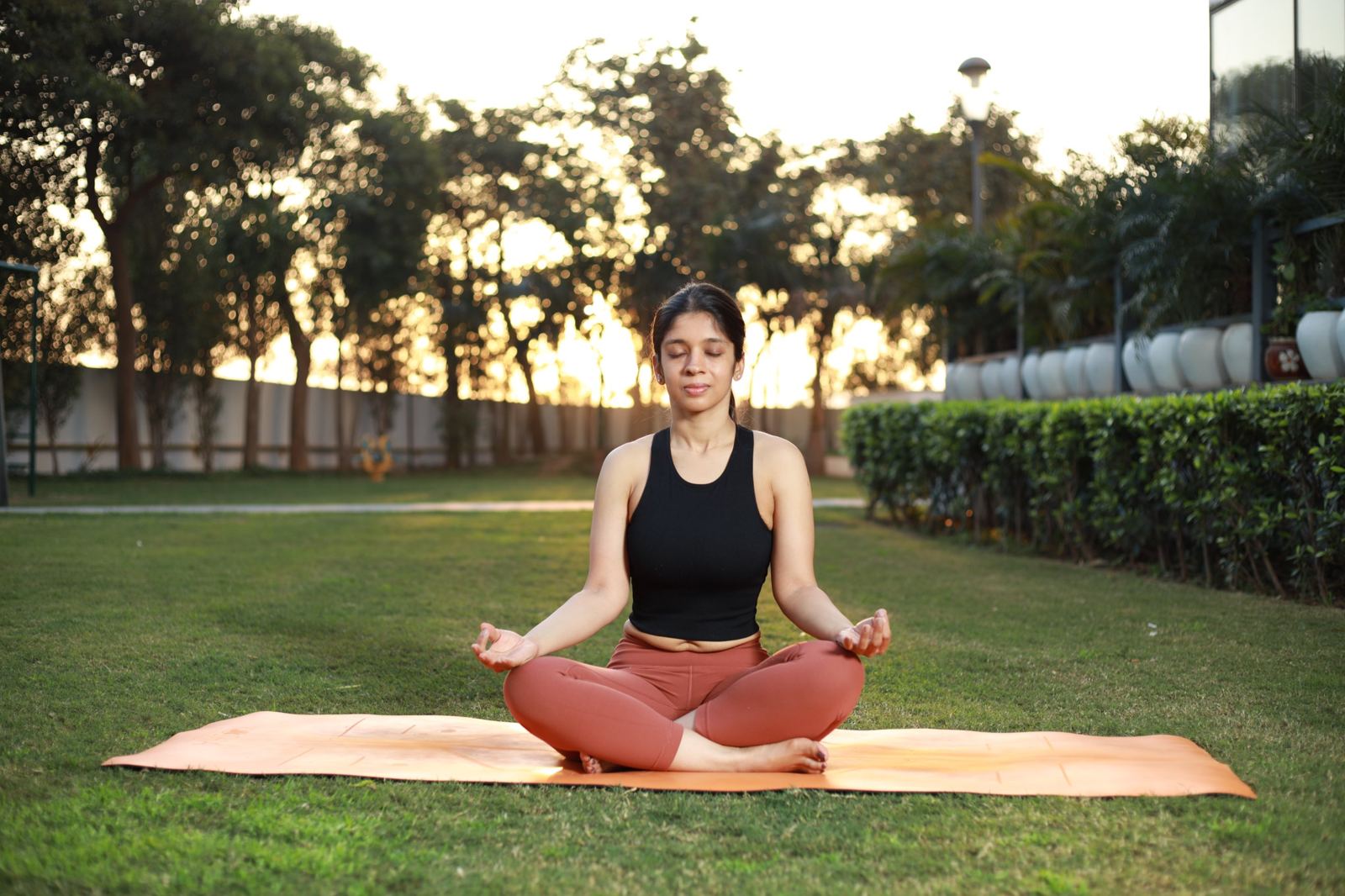 Yoga with Juhie website: Juhie meditating cross-legged on a mat in a garden at sunset